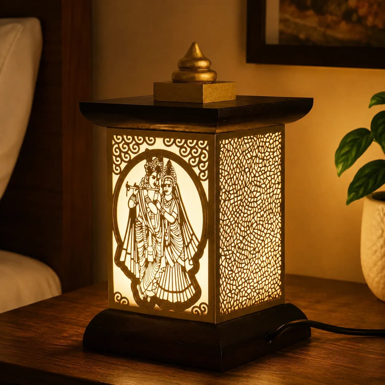 A wooden jharokha lamp with a brass Radha-Krishna filigree panel, lit on a bedside table