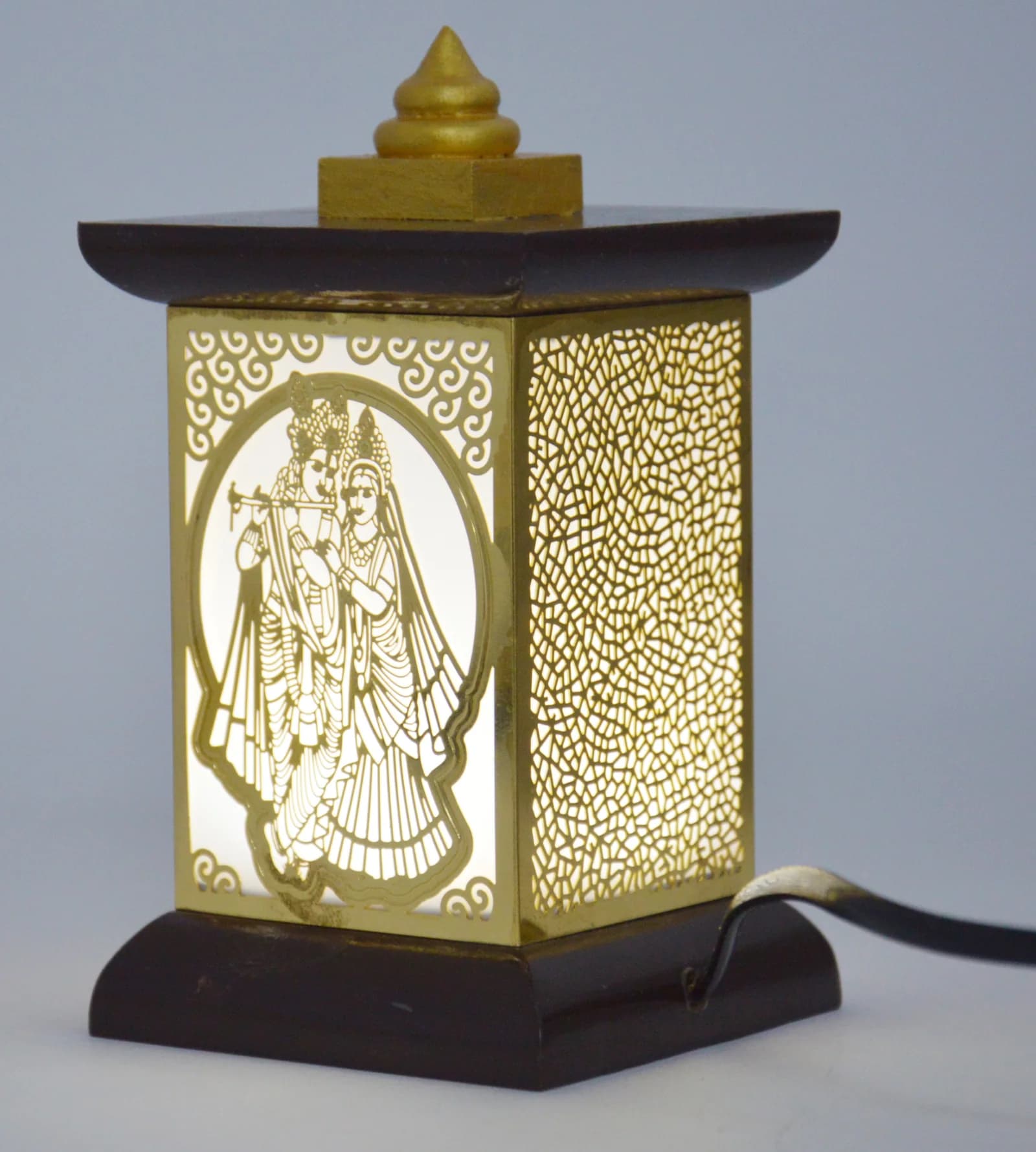 Wooden jharokha table lamp lit from within, its brass Radha-Krishna filigree panel glowing